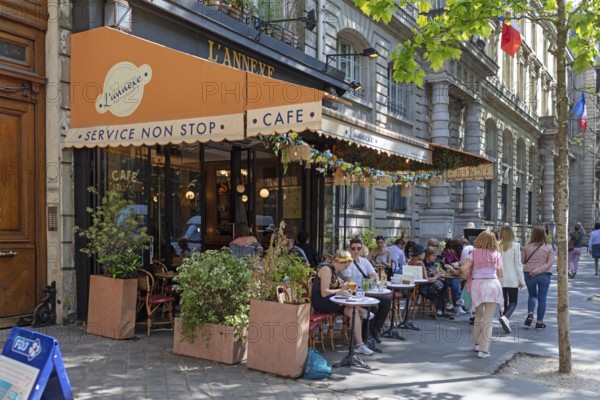 Street café with people sitting at tables, surrounded by plants and an awning, Paris