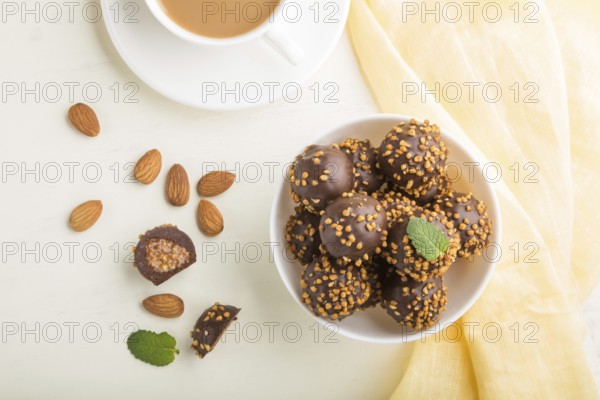 Chocolate caramel ball candies with almonds and a cup of coffee on a white wooden background and yellow textile. top view, flat lay, close up