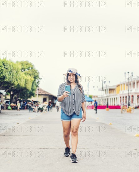 Smiling young tourist in hat walking with cell phone on the street. Female tourist texting phone in the square of Granada