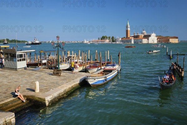 VENICE, ITALY, JULY 19, 2019: Gondolier with client tourists in gondola in lagoon of Venice by Saint Mark (San Marco) square with San Giorgio di Maggiore church seen from Ponte della Paglia in Venice, Italy, Europe