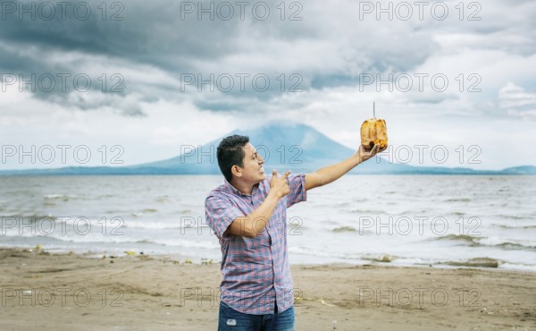 Young man vacationing and enjoying a coconut on the beach. Smiling man holding and pointing at a coconut on a beach in Nicaragua. Young man pointing at a coconut on the beach