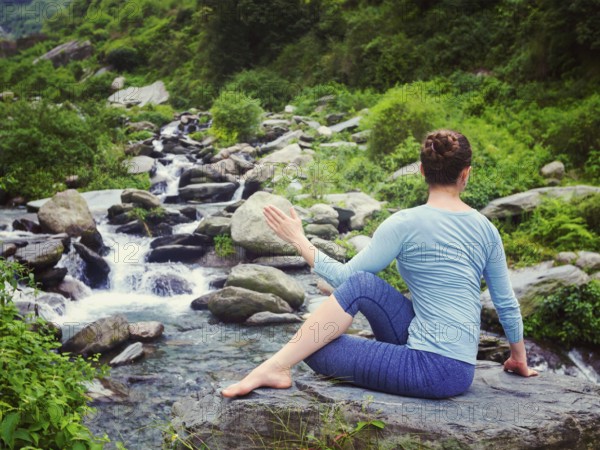 Yoga exercise outdoors, woman doing Ardha matsyendrasana asana, half spinal twist pose at tropical waterfall in Himalayas in India. Vintage retro effect filtered hipster style image