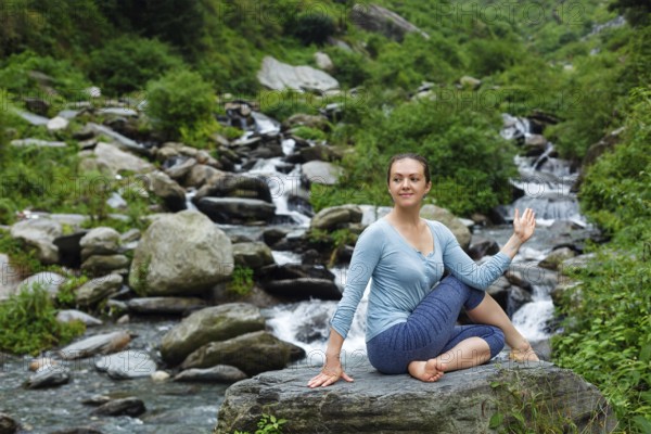 Yoga exercise outdoors, woman doing Ardha matsyendrasana asana, half spinal twist pose at tropical waterfall in Himalayas in India