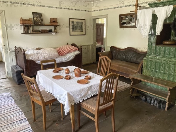 Room with dining table simple earthenware dishes four chairs bed leather sofa tiled stove in teacher's flat in historic rebuilt schoolhouse and town hall from 1830 in Freilichtmuseum Neuhausen, Neuhausen ob Eck, Kreis Tuttlingen, Baden-Württemberg, Germany, Europe