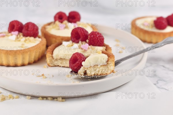 Piece of small tartlet pastry with white cream, topped with raspberry fruits and almond sprinkles on spoon