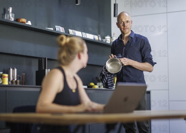 Symbolic photo on the subject of division of labour for couples in the household. A woman sits at a laptop in a kitchen while a man washes dishes in the background. Berlin, 13.08.2024