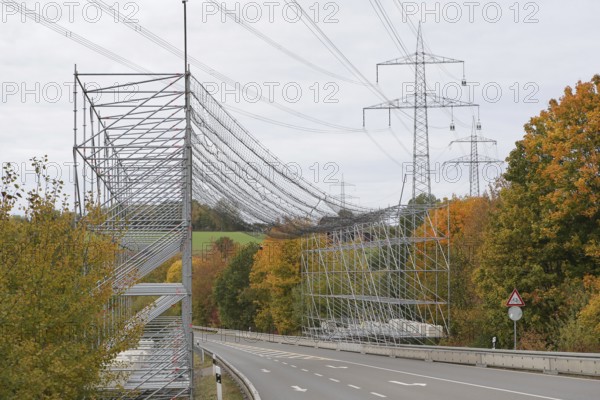 A scaffold with steel net as protection over a road, safety measure due to work on an overhead line and electricity pylons, in the background trees in autumn