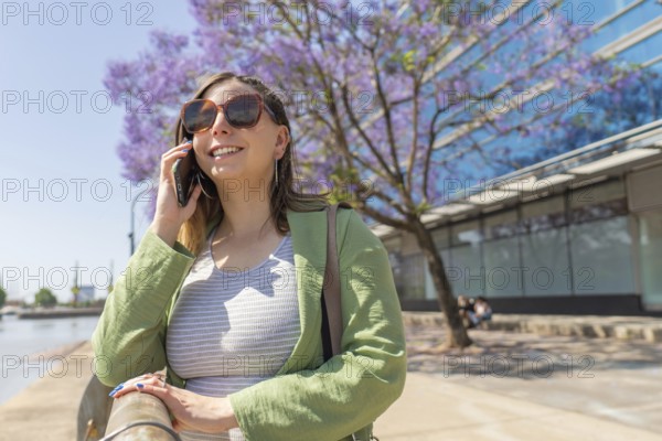 Businesswoman using smartphone near blooming jacaranda mimosifolia tree, enjoying sunny day in urban environment