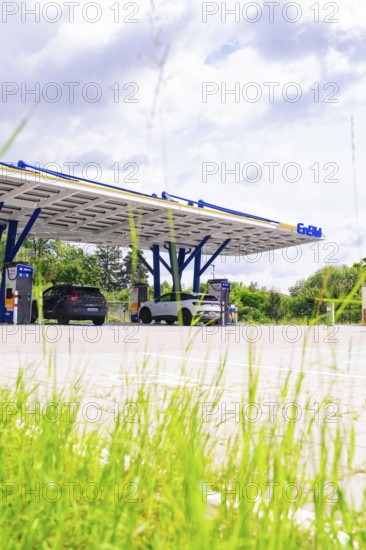 Close-up of grass, behind it EnBW charging stations with solar-powered roof and electric cars, ENBW charging park, Rutesheim, Germany, Europe