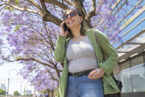 A young woman in casual attire talks on her smartphone while sightseeing by jacaranda trees and a city waterfront. Clear blue sky and urban architecture complete the scenic setting