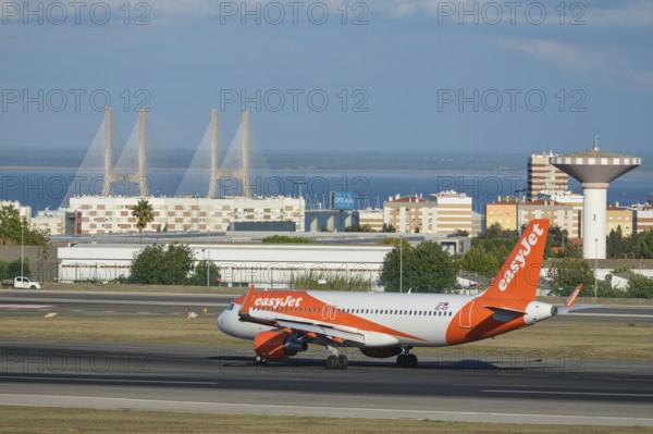 Lisbon, Portugal - September 2, 2023: easyJet Airbus A320-214 passenger plane taxi on runway in Humberto Delgado Airport in Lisbon with Vasco da Gamma bridge in background