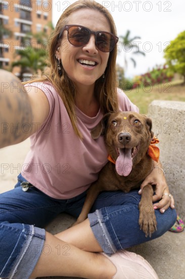 A smiling woman wearing sunglasses sits on the ground, taking a selfie while petting her dog, who looks at her lovingly