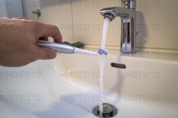 A person holds an electric toothbrush under a running tap in the bathroom, symbolic image of water consumption