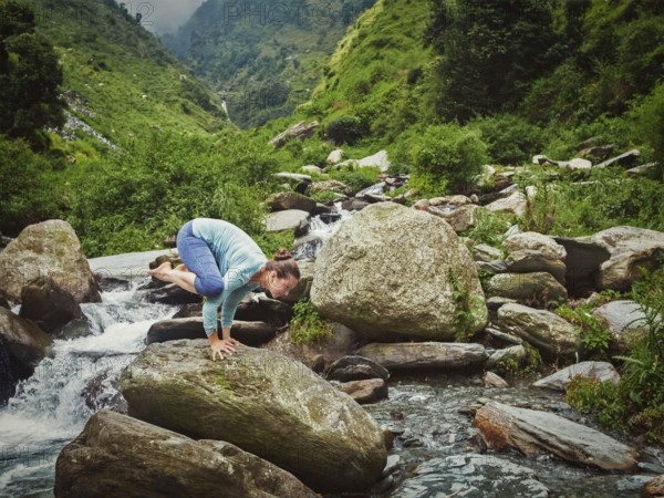 Woman doing Kakasana asana, crow pose arm balance outdoors at tropical waterfall. Vintage retro effect filtered hipster style image