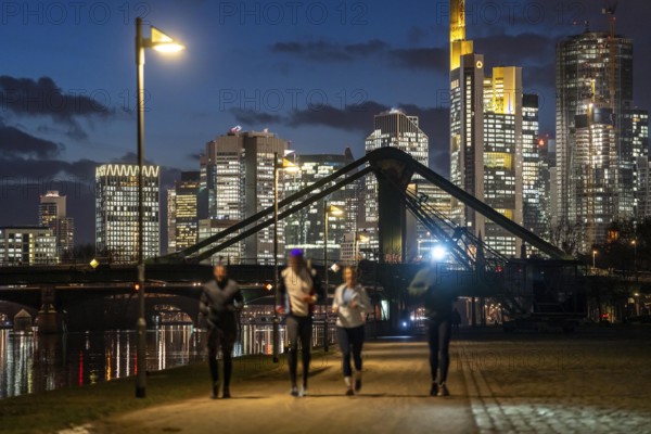 Skyline of the city centre of Frankfurt am Main, joggers on the pavement, promenade along the river Main, dusk, Hesse, Germany raft bridge in the background