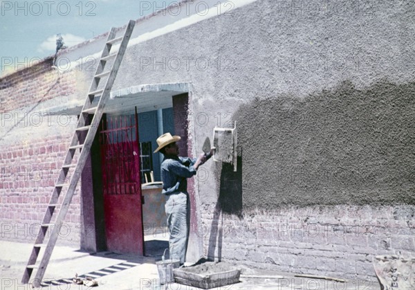 Construction worker throwing cement through a sieve to make wall panelling over modern red bricks, Mexico around 1961