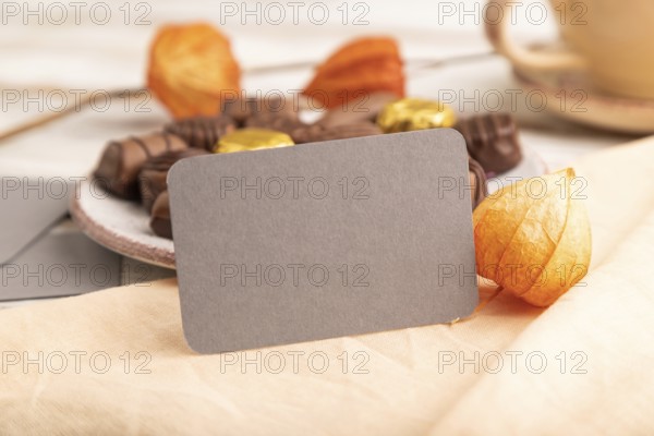 Gray business card with chocolate candies and physalis flowers on a white wooden background and orange textile. side view, close up, selective focus