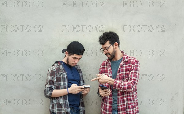 Two young friends leaning against a wall checking their cell phones, Two friends leaning against a wall looking at the content on their phones. Friend showing cell phone to his friend outdoors