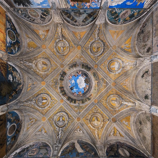 Ducal Palace: vault of the “Bridal Chamber or Camera Picta", which is a room located in the north-east tower of the Castle of San Giorgio in Mantua. It is famous for the cycle of frescoes that covers its walls, a masterpiece by Andrea Mantegna, created between 1465 and 1474.
Detail of the vault illusionistically divided into painted rib vault and pendentives. Fake ribs divide the space into regular figures, with a golden background and monochrome paintings.
In the center is the famous oculus. It is a round open illusionistically towards the sky, which was supposed to recall the famous oculus of the Pantheon. In the oculus you can see a balustrade from which a court lady leans out, accompanied by her black servant, a group of maids, a dozen putti, a peacock and a vase, against the background of a blue sky. To reinforce the impression of the open oculus, Mantegna painted some cherubs dangerously poised clinging to the inside of the frame, with dizzying glimpses of their plump little bo...