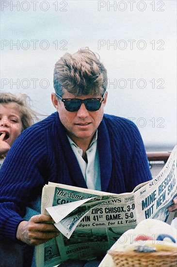 U.S. President John F. Kennedy reading newspaper with  his daughter, Caroline Kennedy in background, aboard the Presidential yacht, “Honey Fitz,” off the coast of Hyannis Port, Massachusetts, USA, Cecil Stoughton, White House Photographs, August 25, 1963