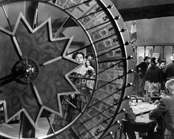 Yvonne De Carlo (viewed through wheel spokes), on-set of the western film, "Border River", Universal Pictures, 1954