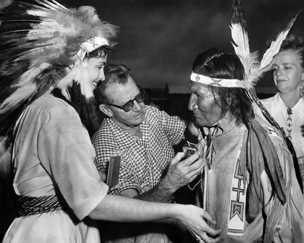 Suzan Ball (left), John Sitting Bull being given a hearing aid (left), on-set of the western film, "Chief Crazy Horse", Universal Pictures, 1955