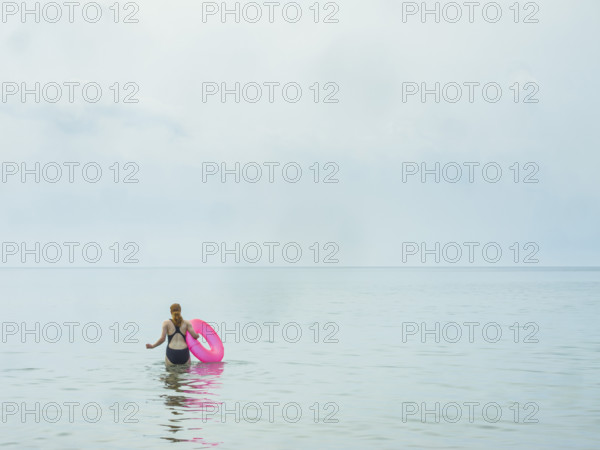 Rear view of young adult woman wading in water holding a pink inner tube