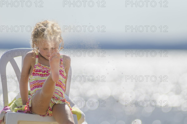 Young girl kicking up sand with her feet while sitting in chair at beach