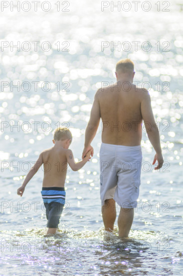 Rear view of father and son holding hands while wading into water at beach