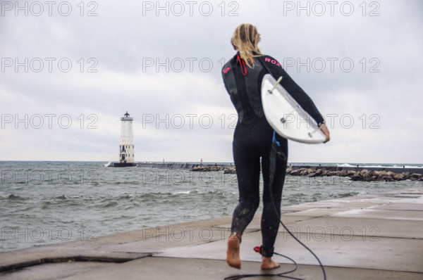 Rear view of young adult woman in wetsuit standing with surf board on Frankfort North lighthouse and pier, Lake Michigan, Frankfort, Michigan, USA