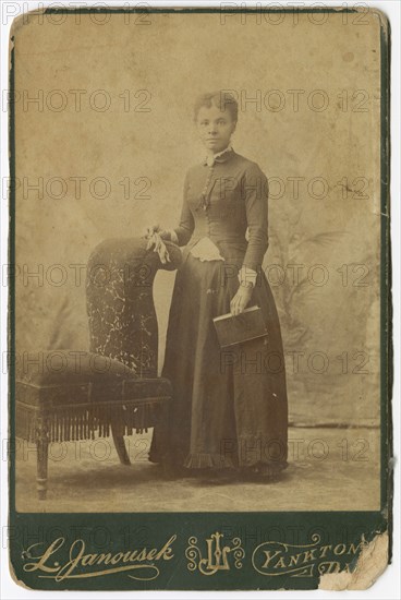 Photograph of a woman standing next to a chair and holding a book, late 19th century. Creator: Louis Janousek.