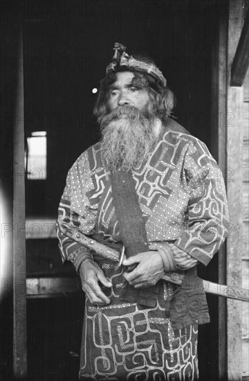 Ainu chief wearing a headdress and holding a sword standing in a doorway, 1908. Creator: Arnold Genthe.