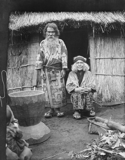 Ainu man and seated woman at the entrance of a hut, 1908. Creator: Arnold Genthe.