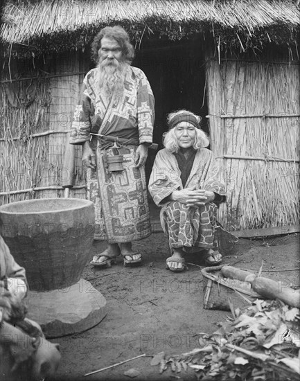 Ainu man and seated woman at the entrance of a hut, 1908. Creator: Arnold Genthe.