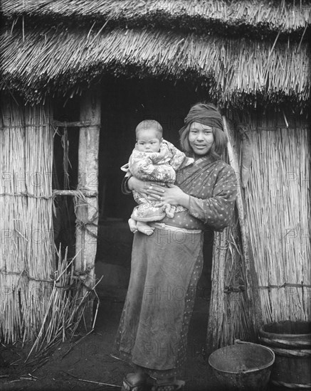 Ainu woman holding a child standing outside a hut, 1908. Creator: Arnold Genthe.