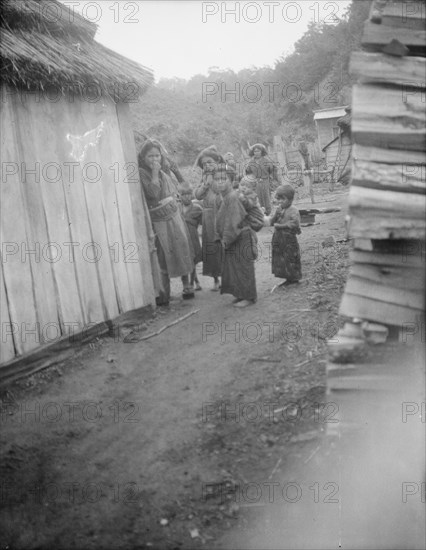 Group of Ainu children standing in a passageway between huts, 1908. Creator: Arnold Genthe.