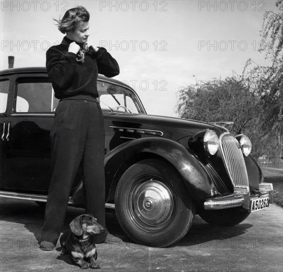 Photographer Kerstin Bernhard stands next to a car with her dachshund, 1955-1960. Creator: Unknown.