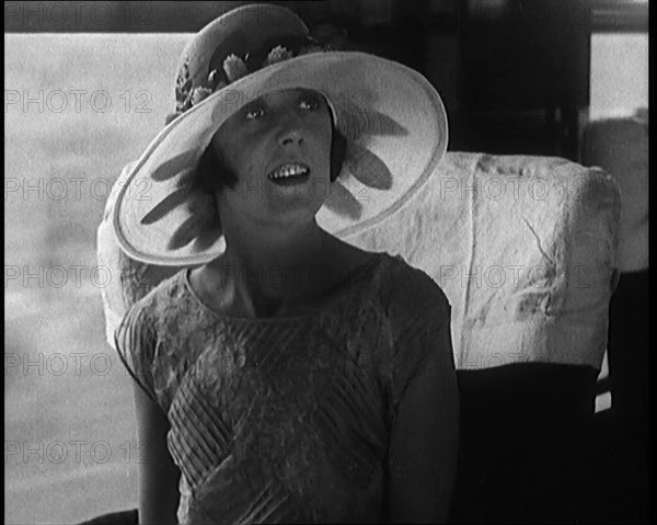 Female Civilian Wearing a Large Fashionable Hat Decorated with Flowers Seating at the Window...1920. Creator: British Pathe Ltd.