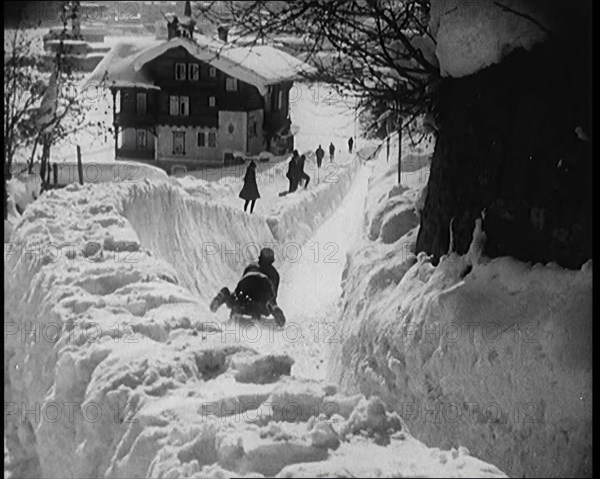 A Female Civilian Sledging down a Snowy Track Watched by Other Civilians with a Picturesque..., 1920 Creator: British Pathe Ltd.