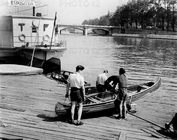 En juin 1950, à Paris, sur les bords de Seine, dans le port de plaisance, deux adolescents mettent un canoë en bois à l'eau, aidés par deux employés du TOURING-CLUB-DE-FRANCE.  Cette association, fondée en 1890 par Edouard BRUEL, a pour but de développer le tourisme, d'aménager des espaces de loisirs et de détente, d'inventorier et de sauvegarder les sites naturels.