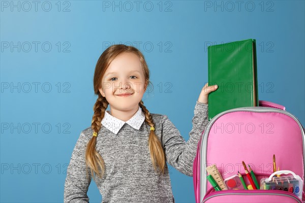 Closeup portrait of little girl schoolgirl on blue background. The child puts in a satchel book. Back to school. The concept of education.