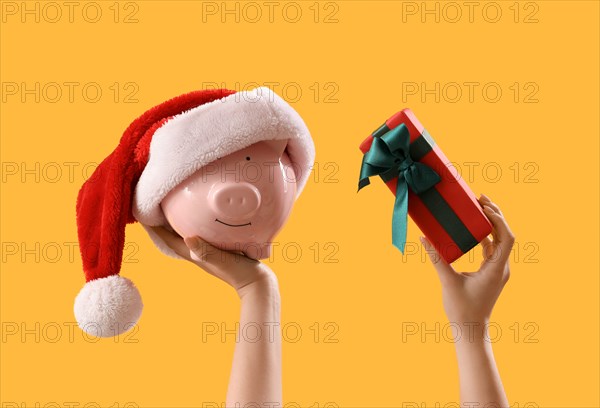 Hands holding Christmas gift, piggy bank and Santa Claus hat on yellow background