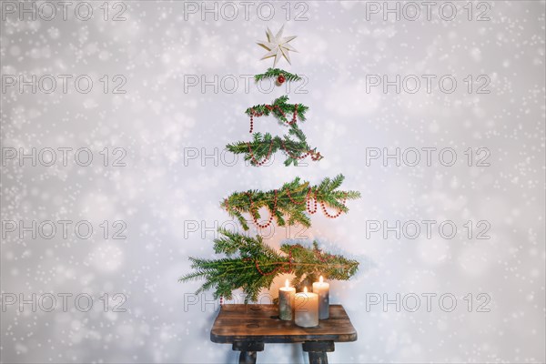 Christmas tree alternative, evergreen branches with decoration hanging on the wall above a rustic wooden stool with candles, sus