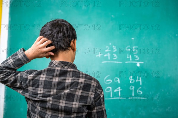 Confused kid scratching head to solve mathematics problem on board at classroom - concept of intelligence, trouble learning and education