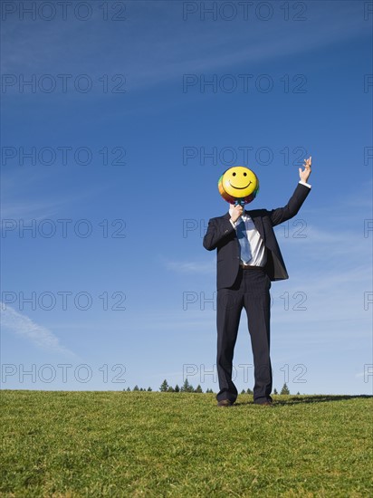 Businessman holding smiley face balloon over face.