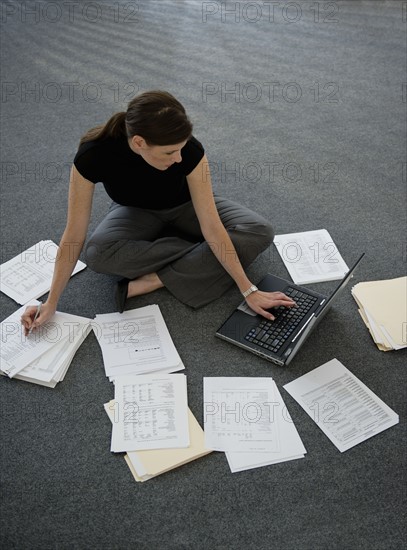 Woman sitting on floor with laptop with documents around.