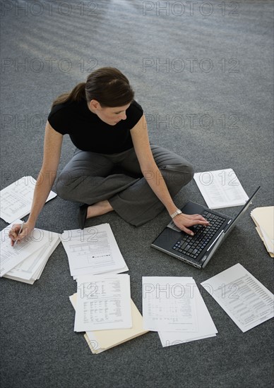 Woman sitting on floor with laptop with documents around.