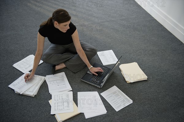 Woman sitting on floor with laptop with documents around.