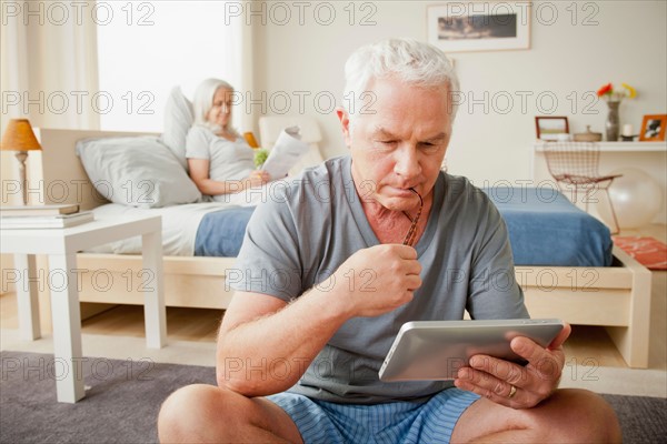 Senior man holding photo frame, woman sitting on bed in background. Photo : Rob Lewine
