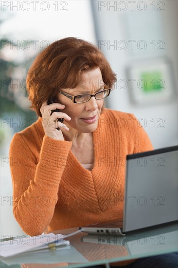 Senior woman working on laptop and talking on cell phone.